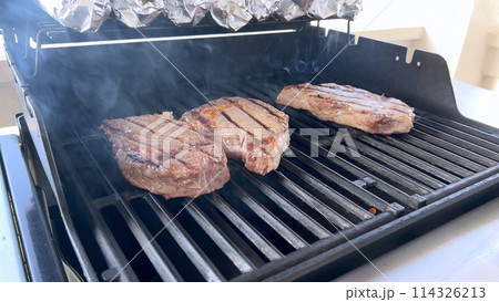 A person uses tongs to flip juicy steaks on a grill, with flames licking the meat, captured in a moment of intense heat and sizzle, showcasing a classic outdoor cooking experience. 114326213