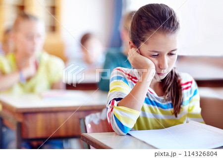 Child, school and desk with book or bored in classroom or education lesson or reading, knowledge or studying. Girl, kids and unhappy or learning or tired pupil with paper or fatigue, student or moody 114326804