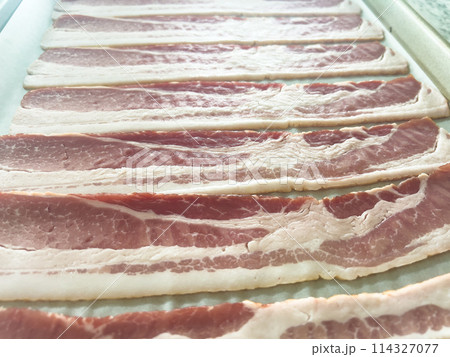 Neatly arranged raw bacon strips on a baking tray, prepared for cooking, capturing the fresh, uncooked look of this popular breakfast ingredient before it turns crispy and golden. Neatly arranged raw bacon strips on a baking tray, prepared for cooking, capturing the fresh, uncooked look of this popular breakfast ingredient before it turns crispy and golden. 114327077
