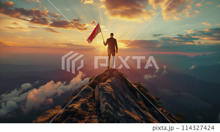 A man is standing on a mountain top holding a flag. The flag is red, white, and blue. The sky is cloudy and the sun is setting. The man is looking up at the flag, as if he is about to fly it 114327424