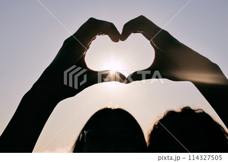 Silhouette, heart hands and couple at sky for sunset, love and care together on summer holiday with flare. Sign, closeup and romance at beach with fingers for peace, support and connection emoji 114327505