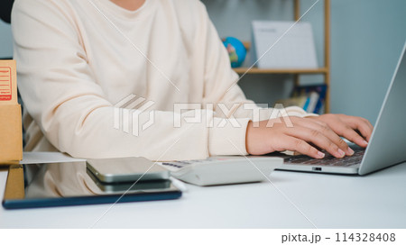 Woman runs an e-commerce business is checking orders from laptop, she owns an online store, she packs and ships through a private transport company. Online selling and online shopping concepts. Woman runs an e-commerce business is checking orders from laptop, she owns an online store, she packs and ships through a private transport company. Online selling and online shopping concepts. 114328408