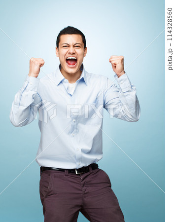 Portrait, screaming and man with surprise, celebration and expression on blue studio background. Face, person and model with achievement, shouting or wow with winner, promotion or giveaway with smile 114328560