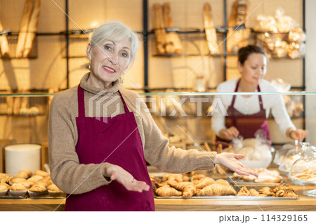 Elderly bakeshop saleswoman in maroon apron recommending baked goods Elderly bakeshop saleswoman in maroon apron recommending baked goods 114329165