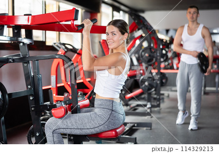 Girl doing strength training in gym, exercising upper body muscles on shoulder press machine Girl doing strength training in gym, exercising upper body muscles on shoulder press machine 114329213