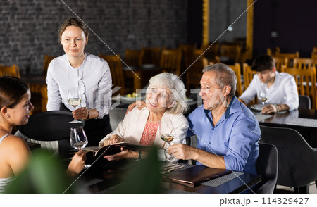Waitress bringing aperitif to elderly couple selecting dishes from menu Waitress bringing aperitif to elderly couple selecting dishes from menu 114329427