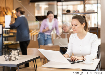 Focused woman drinking coffee while working on laptop in cozy cafe Focused woman drinking coffee while working on laptop in cozy cafe 114329461