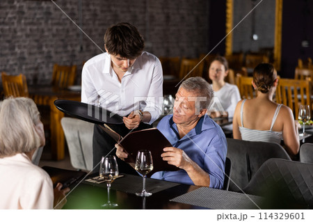 Elderly husband and wife reads menu and makes order to waiter. 114329601