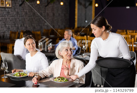 Smiling waitress fulfills order of young girl and elderly woman of restaurant visitors. 114329799