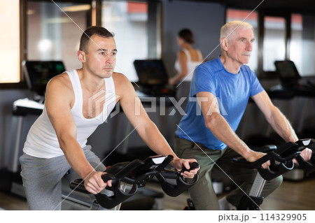 Two men exercising on stationary exercise bikes in gym Two men exercising on stationary exercise bikes in gym 114329905
