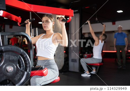Girl doing strength training in gym, exercising upper body muscles on shoulder press machine Girl doing strength training in gym, exercising upper body muscles on shoulder press machine 114329949