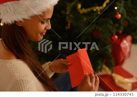 Happy woman, christmas tree and letter with note of gift, love and holiday season in living room. Young person, smile and card for present for xmas wishes, gratitude and kindness in spirit of giving 114330745