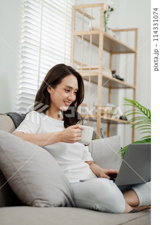 Woman takes pleasure in sipping her coffee as she focuses on her laptop, comfortably seated on a couch in a bright home setting. 114331074