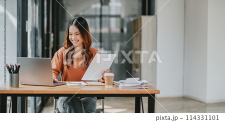 Young businesswoman smiling while examining a document, comfortably working at a spacious office with her laptop. 114331101