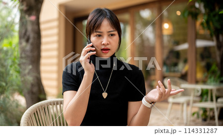 An Asian woman sits at an outdoors table of a cafe having a serious conversation on the phone. 114331287