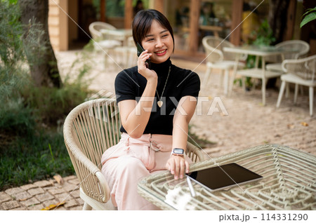 An attractive Asian woman sits at an outdoor table in a garden of a cafe talking on the phone. 114331290