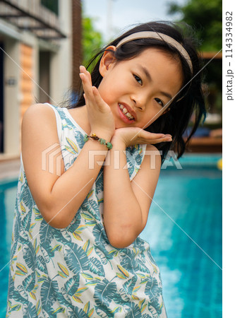 Portrait of an Asian cute girl with cute act in front of a resort swimming pool, girl in casual dress, eyes looking at camera, vertical image. Portrait of an Asian cute girl with cute act in front of a resort swimming pool, girl in casual dress, eyes looking at camera, vertical image. 114334982