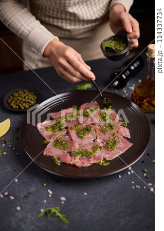tuna carpaccio - woman puts capers green sauce onto slices of fresh raw tuna fillet on black ceramic plate 114337734