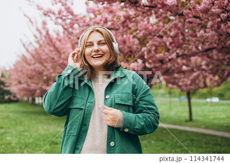 Portrait of charming 30s woman in massive headphones listening to music in the park. Music lover enjoying music. Spring background with a branch of blooming sakura. Tenderness. 114341744