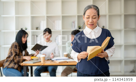 A smiling senior Asian female teacher professor standing in the classroom with a book in her hand. A smiling senior Asian female teacher professor standing in the classroom with a book in her hand. 114341959