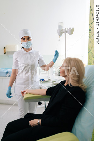 Vertical portrait of nurse in medical uniform preparing and adjusting infusion for woman patient in hospital or beauty salon. Doctor preparing to insert IV needle into patient arm. Healthcare concept. 114342130