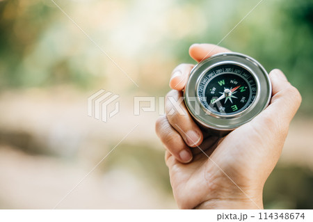 A traveler holding a compass in a park seeks guidance and direction in nature beauty. The compass in the woman hand signifies exploration and a journey to find one path. 114348674