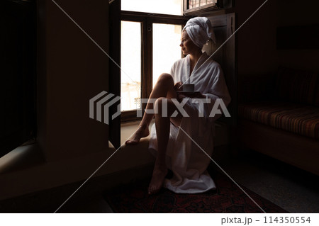 Woman in dark room sitting in robe by window. Attractive young girl in bathrobe and with towel on her head is holding cup, looking at window and smiling while sitting on window sill in old hotel room 114350554