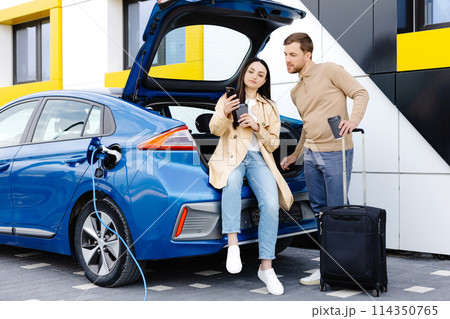 Young friends charging automobile battery from public station and standing near parking for electric car. Man and woman talking and drinking coffee in city Young friends charging automobile battery from public station and standing near parking for electric car. Man and woman talking and drinking coffee in city 114350765