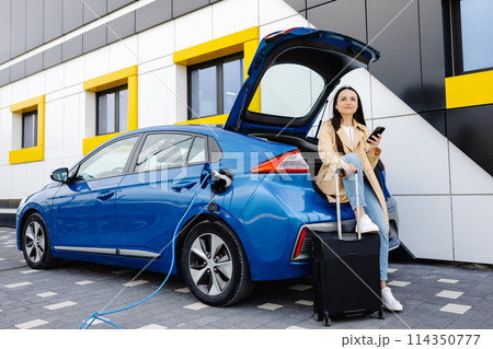 Young woman waiting charging automobile battery from small public station and using smartphone while charging automobile 114350777