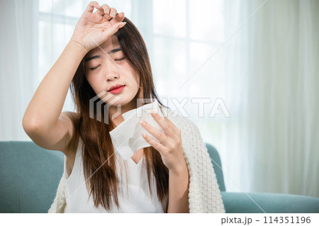 Sick woman sitting under blanket on sofa and sneeze with tissue paper in living room, Asian young female blowing nose sneezing in tissue at home, fever caught cold Sick woman sitting under blanket on sofa and sneeze with tissue paper in living room, Asian young female blowing nose sneezing in tissue at home, fever caught cold 114351196
