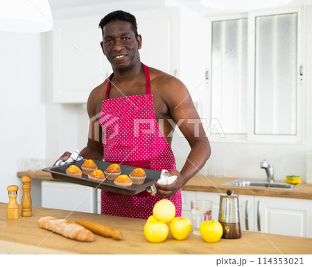 Smiling man with naked torso in an apron holding sheet pan with just baked cupcakes in kitchen Smiling man with naked torso in an apron holding sheet pan with just baked cupcakes in kitchen 114353021
