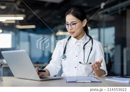 Professional Asian female doctor in a white coat engaged in a telemedicine consultation, using a laptop and speaking expressively in a modern office setting. 114353022