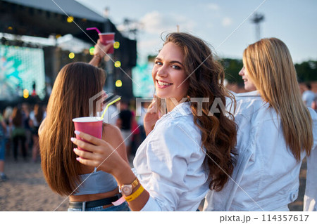 Group of girls enjoy music at a beach festival, smiling with drinks in hand, sun setting. Casual summer party vibe, DJ on stage, crowd dancing, fun vacation moment by the sea. Group of girls enjoy music at a beach festival, smiling with drinks in hand, sun setting. Casual summer party vibe, DJ on stage, crowd dancing, fun vacation moment by the sea. 114357617