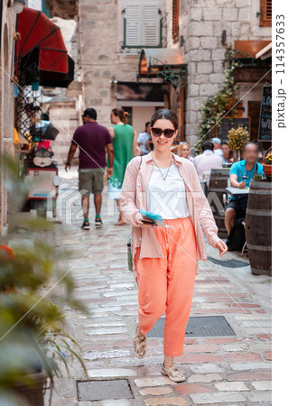 Young Caucasian woman holds a paper map in her hand and walks around an old European city for sightseeing. Summer vacation alone. Vertical orientation 114357633