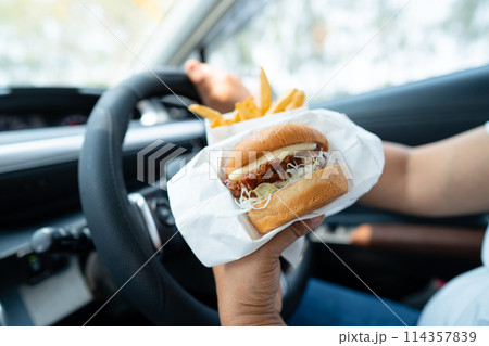 Asian lady holding hamburger and French fries to eat in car, dangerous and risk an accident. 114357839