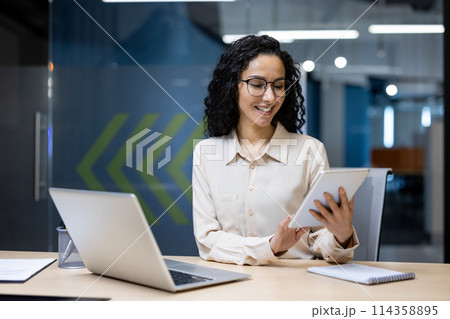 Young successful businesswoman at workplace is satisfied with results of achievement at work smiling and using tablet computer, programmer is testing new software in application Young successful businesswoman at workplace is satisfied with results of achievement at work smiling and using tablet computer, programmer is testing new software in application 114358895