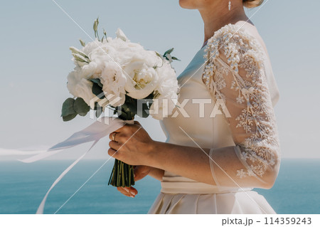 bride is holding a bouquet of white flowers. The bouquet is large and has a lot of flowers in it. The woman is wearing a white dress and a gold ring on her finger. Scene is elegant and romantic. 114359243
