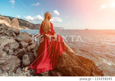 Red dress sea woman. A blonde with flowing hair in a long red dress sits on a rock near the sea. The concept of trips, a photo shoot at the sea 114359245