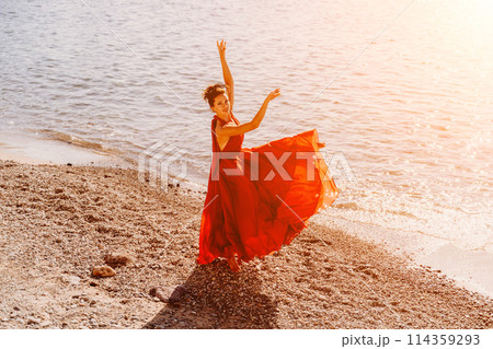 Woman red dress sea. Female dancer in a long red dress posing on a beach with rocks on sunny day. Girl on the nature on blue sky background. Woman red dress sea. Female dancer in a long red dress posing on a beach with rocks on sunny day. Girl on the nature on blue sky background. 114359293