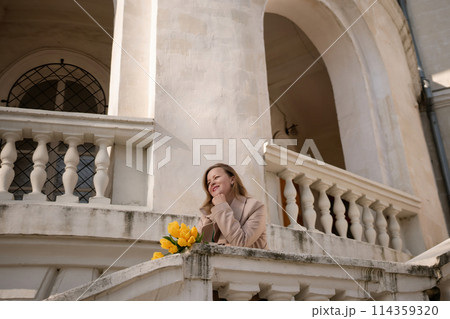 A woman wearing sunglasses and holding a bouquet of yellow flowers stands on a balcony. The scene is peaceful and serene, with the woman looking out over the city. 114359320