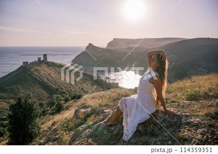 A woman in a white dress is sitting on a rock overlooking a body of water. She is enjoying the view and taking in the scenery. A woman in a white dress is sitting on a rock overlooking a body of water. She is enjoying the view and taking in the scenery. 114359351