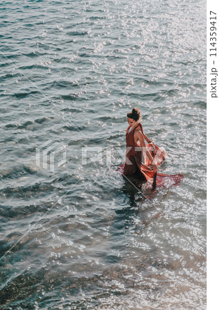 Woman red dress sea. Female dancer in a long red dress posing on a beach with rocks on sunny day Woman red dress sea. Female dancer in a long red dress posing on a beach with rocks on sunny day 114359417