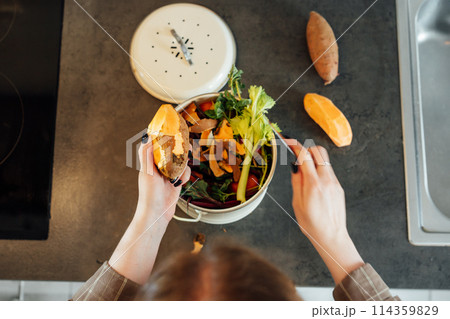 Compost the kitchen waste, recycling at home. Woman putting vegetables cutted leftovers into the garbage, compost bin on the table of kitchen. Environmentally responsible, ecology. Selective focus. 114359829