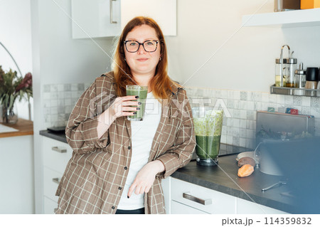 Smiling plus size young woman with just made glass of detox shake, green smoothie in the kitchen. Healthy dieting, eating, cooking, slimming. Natural weight loss program. Vegan, vegetarian diet. 114359832