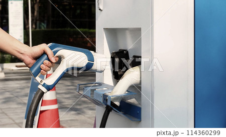 Driver of electric car hold batteries charger plug. Hand holding EV charging plug in front of camera with blurred background of outdoor. Sustainable energy powered electric charging station background Driver of electric car hold batteries charger plug. Hand holding EV charging plug in front of camera with blurred background of outdoor. Sustainable energy powered electric charging station background 114360299