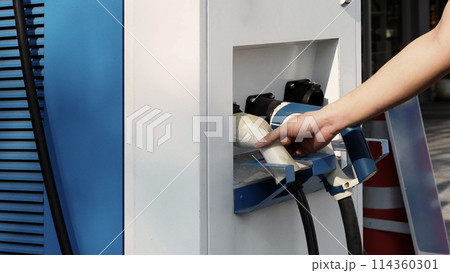 Driver of electric car hold batteries charger plug. Hand holding EV charging plug in front of camera with blurred background of outdoor. Sustainable energy powered electric charging station background Driver of electric car hold batteries charger plug. Hand holding EV charging plug in front of camera with blurred background of outdoor. Sustainable energy powered electric charging station background 114360301