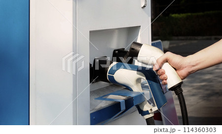 Driver of electric car hold batteries charger plug. Hand holding EV charging plug in front of camera with blurred background of outdoor. Sustainable energy powered electric charging station background Driver of electric car hold batteries charger plug. Hand holding EV charging plug in front of camera with blurred background of outdoor. Sustainable energy powered electric charging station background 114360303