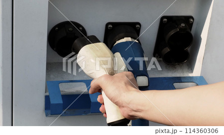 Driver of electric car hold batteries charger plug. Hand holding EV charging plug in front of camera with blurred background of outdoor. Sustainable energy powered electric charging station background Driver of electric car hold batteries charger plug. Hand holding EV charging plug in front of camera with blurred background of outdoor. Sustainable energy powered electric charging station background 114360306