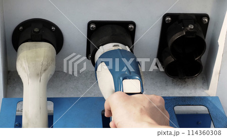 Driver of electric car hold batteries charger plug. Hand holding EV charging plug in front of camera with blurred background of outdoor. Sustainable energy powered electric charging station background Driver of electric car hold batteries charger plug. Hand holding EV charging plug in front of camera with blurred background of outdoor. Sustainable energy powered electric charging station background 114360308