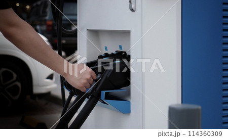 Driver of electric car hold batteries charger plug. Hand holding EV charging plug in front of camera with blurred background of outdoor. Sustainable energy powered electric charging station background Driver of electric car hold batteries charger plug. Hand holding EV charging plug in front of camera with blurred background of outdoor. Sustainable energy powered electric charging station background 114360309